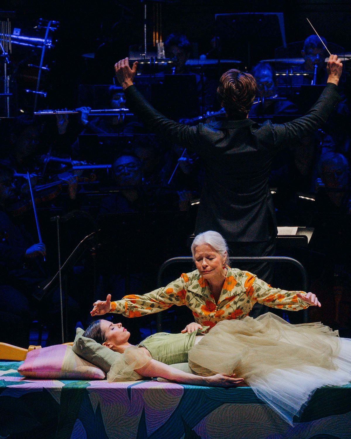 The conductor raises his arms above a sleeping princess and an old woman spreading her arms.