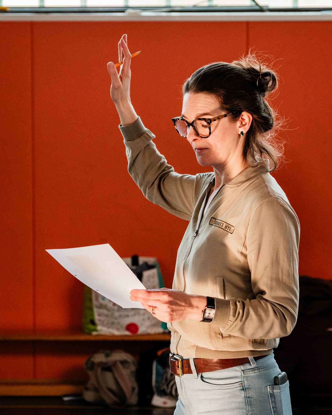 Anisha Bondy raises her arm at a rehearsal, a sheet of paper in her hand, in front of an orange wall.